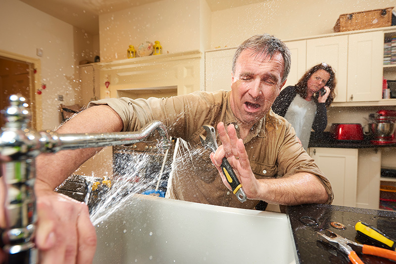 man trying to fix sink unsuccessfully with woman calling a professional in the background