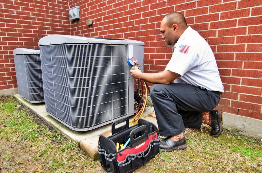 Man in white uniform crouched beside an outside AC unit performing AC maintenance