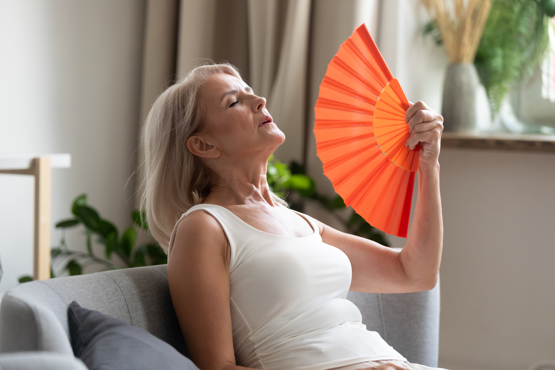 woman in white tank top fanning herself with orange handheld fan looks tired and hot in her home